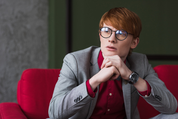 Male fashion model in eyeglasses dressed in suit sitting on red couch - Fotografie, Obrázek