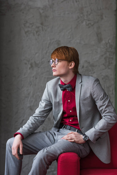 Profile of male model in eyeglasses dressed in suit sitting on edge of couch - 写真・画像