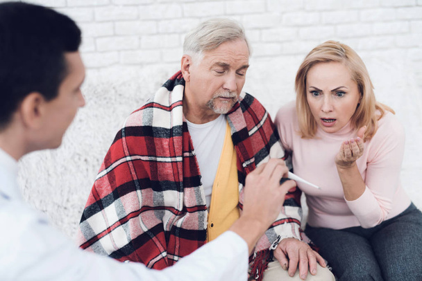 A doctor came to the old man in a yellow cardigan. The doctor shows the thermometer, the woman is in shock. They are in the house of an elderly couple. - Photo, image