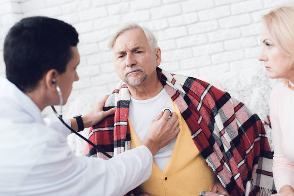 A doctor came to the old man in a yellow cardigan. The old man became ill and the doctor listens to his heart with a stethoscope. Next to him is the old man's wife. - Photo, image