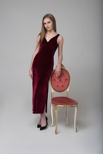 Young long-haired girl poses in long red dress near chair - Foto, imagen