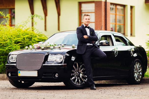 the fiance in a black suit stands near a beautiful black car - Foto, Bild