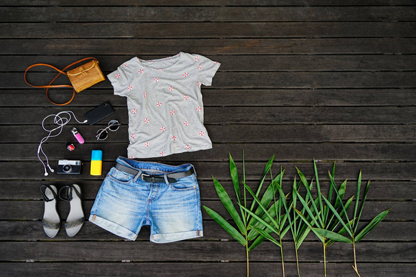 Woman summer clothes collage flat lay isolated on dark wood background. Feminine desk top view fashion accessories: t-shirt, shorts, sandals, sunglasses, vintage photo camera, headphones. Holiday - Photo, Image