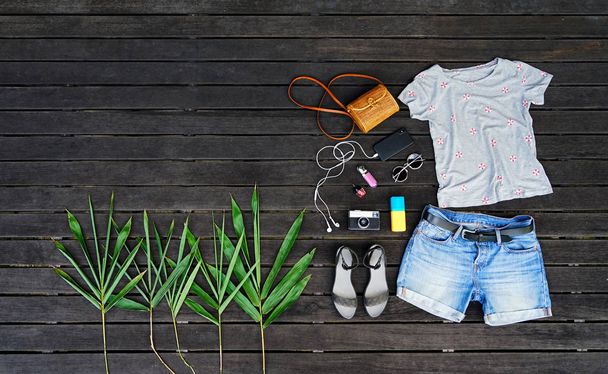 Woman summer clothes collage flat lay isolated on dark wood background. Feminine desk top view fashion accessories: t-shirt, shorts, sandals, sunglasses, vintage photo camera, headphones. Holiday - Photo, Image