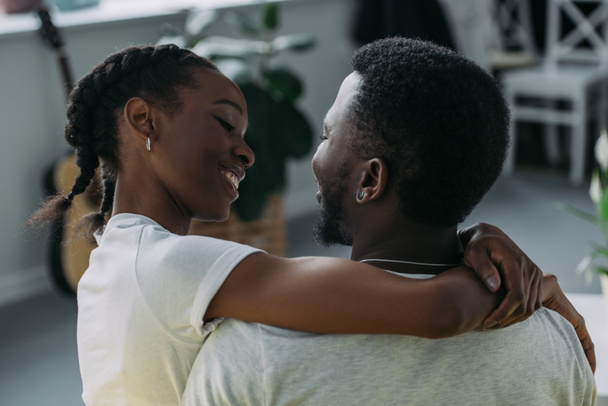 zadní pohled na mladé afroamerické pár všeobjímající a usmívá se navzájem doma - Fotografie, Obrázek