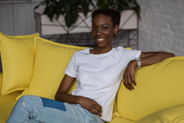 beautiful african american girl sitting on yellow couch and smiling at camera - Фото, изображение