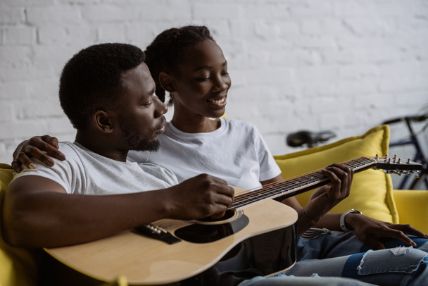 happy young african american couple sitting together on sofa and man playing acoustic guitar - Foto, Imagem