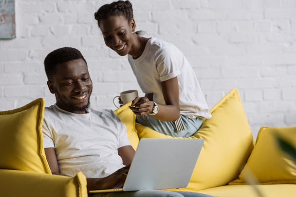 smiling young african american couple using laptop while sitting on sofa at home  - Foto, immagini
