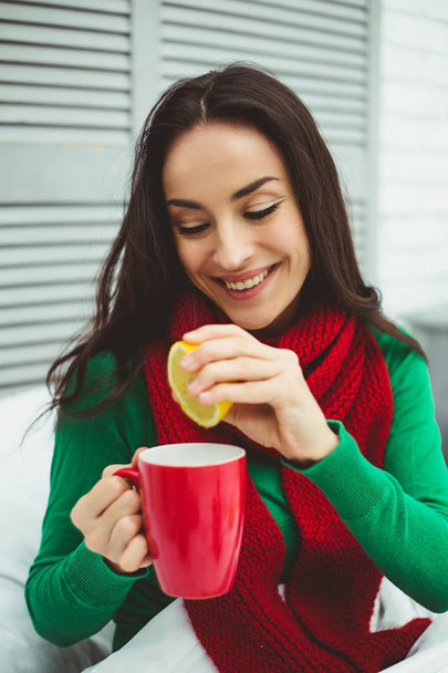No more coughing. A young beautiful smiling woman in a red scarf is cured for cough, drinks hot tea with a lemon at home on the bed. - Photo, Image