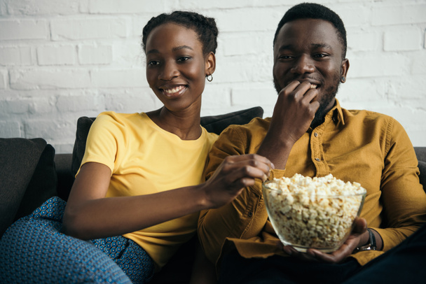 Lächelndes junges afrikanisch-amerikanisches Paar isst Popcorn, während es zu Hause auf dem Sofa sitzt  - Foto, Bild