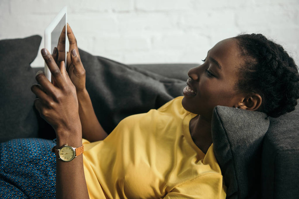 自宅のソファの上に横たわっている間デジタル タブレットを使用して笑顔の若いアフリカ系アメリカ人の女性 - 写真・画像