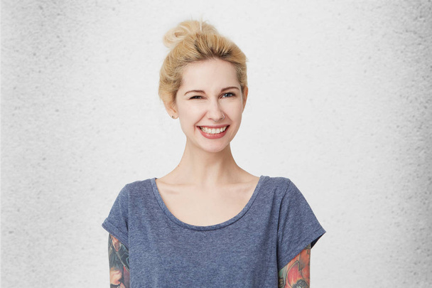Portret młodej dziewczyny Śliczne blond włosy, przebili nos i tatuaże, noszenie na co dzień niebieski t-shirt uśmiechając się i wink. Na białym tle nad studio białe ściany. Koncepcja szczęśliwy - Zdjęcie, obraz