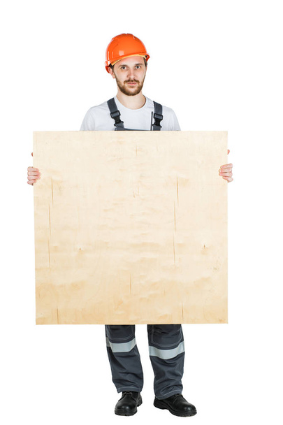 Ένας οικοδόμος ή ένας εργαζόμενος σε ένα προστατευτικό κράνος με ένα φύλλο του κοντραπλακέ. Απομονωμένα σε λευκό φόντο. - Φωτογραφία, εικόνα