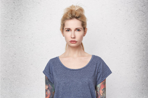 Portrait of serious blonde girl looking directly in camera cross her hands back. Wearing casual blue t-shirt. Tattooes. Pierced nose. Isolated over studio white wall. - Photo, Image