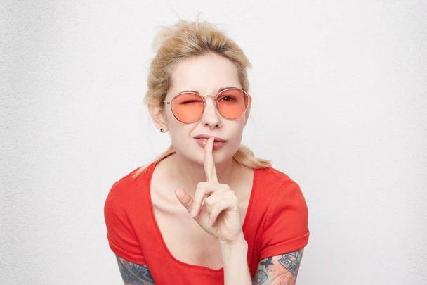 Close-up portret van vrij blond meisje Toon een gebaar van stilte en knipoog. Positieve emotie. Draagt casual rode t-shirt en glazen.  - Foto, afbeelding