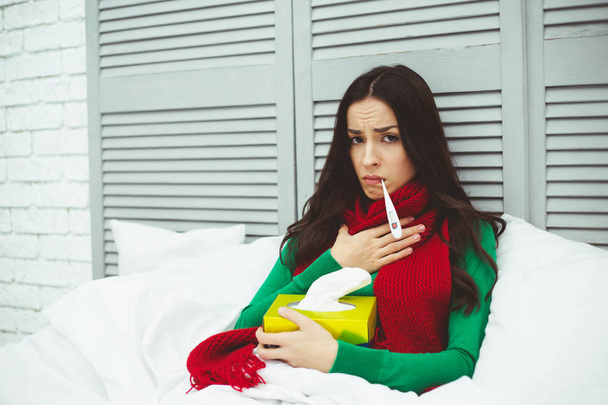 Очень высокая температура тела. Молодая больная женщина в красном шарфе измеряет температуру термометром. Концепция здоровья и болезней
. - Фото, изображение