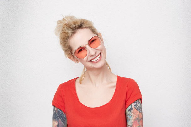 Portret van vrij blond meisje lacht en rechtstreeks in de camera kijken. Casual rode t-shirt en rood stijlvolle bril draagt. Positieve emoties. Doorboorde neus  - Foto, afbeelding