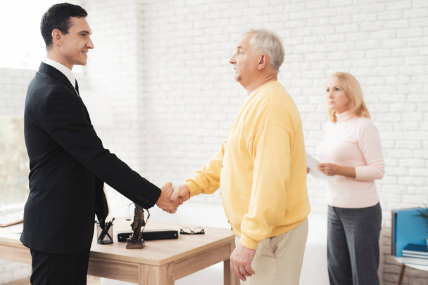 A couple of old people came to see a realtor. An old man in a yellow cardigan greets a realtor in a black suit. Behind him stands a woman in a pink sweater. - Photo, image