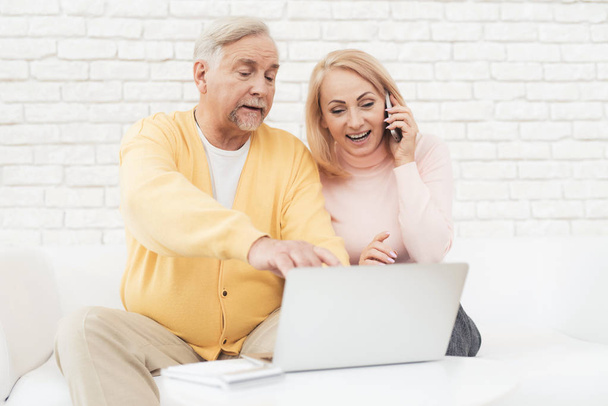 A pair of old people are sitting in front of a laptop. They found the home of their dreams and are happy about it. They are sitting on a large white sofa in their apartment. - Foto, Bild