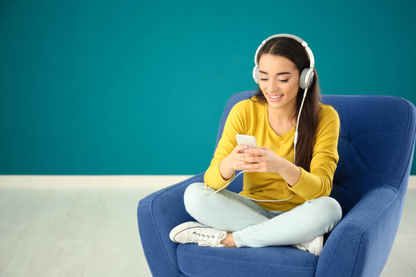 Beautiful young woman listening to music while sitting in comfortable armchair against color wall - Foto, imagen