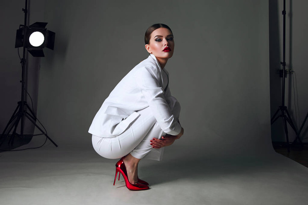 Beautiful young girl with a beautiful figure in a white trouser suit and red shoes sits in a beautiful pose on the floor in the studio. - 写真・画像