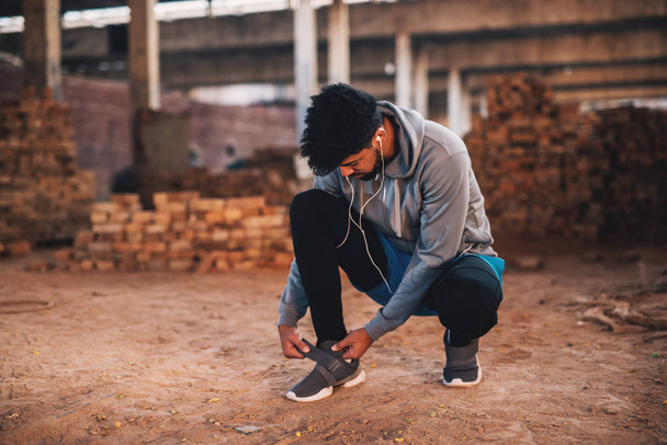  young active male runner in sportswear tying his velcro shoe in abandoned place - Photo, Image