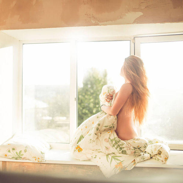 portrait of young naked woman with long red hair on windowsill near windows at morning - Φωτογραφία, εικόνα