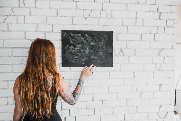 back view of young red hair woman in black dress with black chalkboard on white brick wall background - Foto, Bild