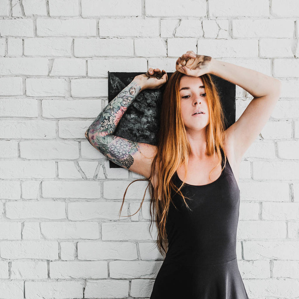 portrait of young red hair woman with tattoo in black dress with black picture on white brick wall background - Photo, Image