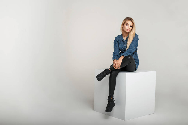 Fashionable young model woman in fashion vintage jeanswear in leather pants and stylish sneakers sits on a white square in the studio - Foto, Imagem