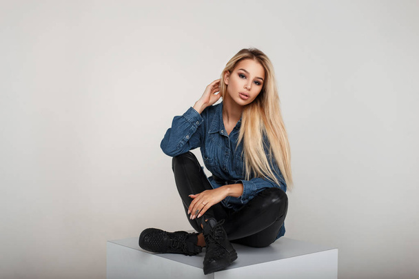 Modieuze jonge blonde meisje in mode denim shirt met zwarte sneakers in studio - Foto, afbeelding
