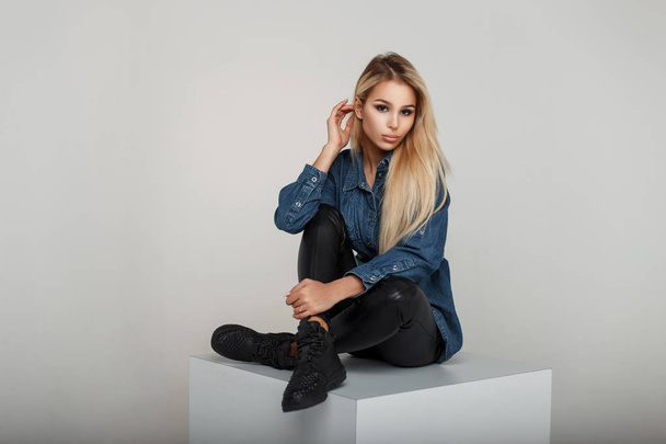 Fashionable young blond woman in fashionable vintage jeanswear with sneakers sits in studio - Fotografie, Obrázek