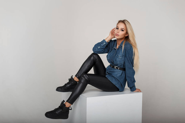 Молода модна блондинка в модній джинсовій сорочці з чорними стильними кросівками позує в студії
 - Фото, зображення