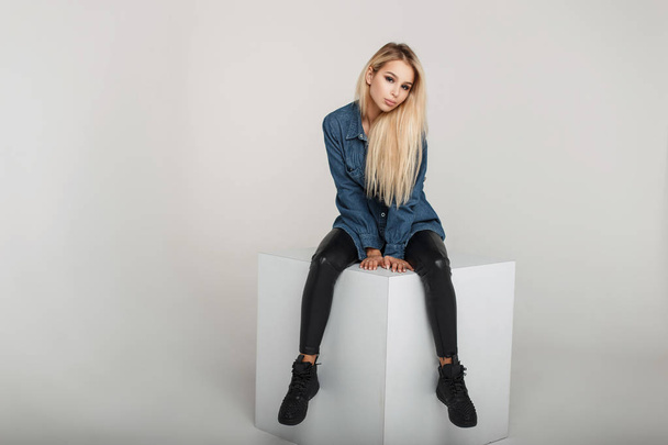 Sexy beautiful young woman in fashionable denim jeans shirt in leather pants and black stylish shoes sits on a white square in the studio - Photo, Image