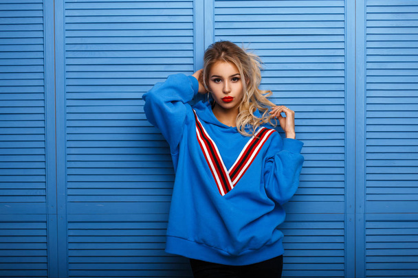 Beautiful young woman with make-up and hairstyle in stylish hoodie poses near blue wooden wall - Photo, Image