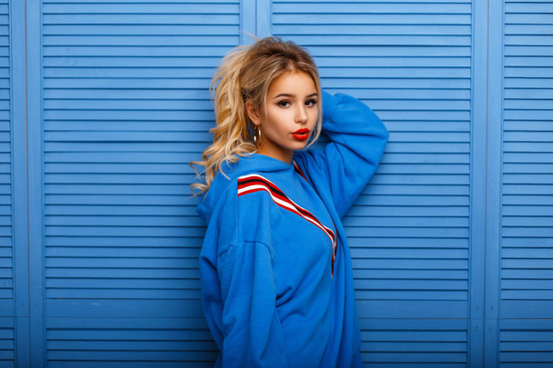 Fashionable model woman with red lips in fashion hoodie posing near blue wooden wall - Foto, immagini