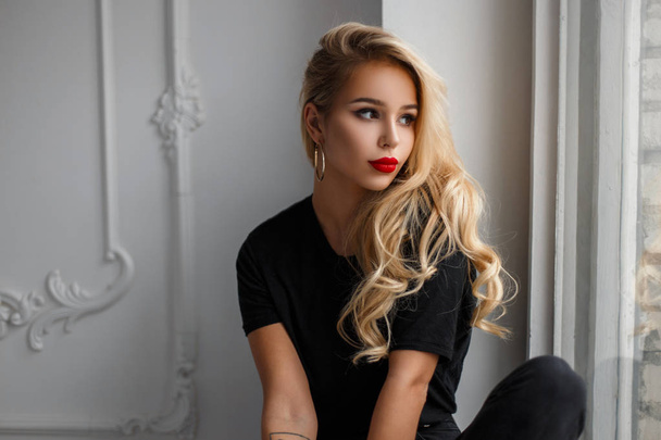 schöne stylische junge Frau in einem modischen schwarzen T-Shirt sitzt und aus dem Fenster schaut - Foto, Bild