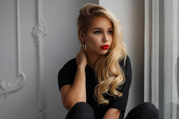 Hermosa joven modelo con estilo chica con labios rojos en una camisa negra de moda sentado cerca de la ventana
 - Foto, imagen