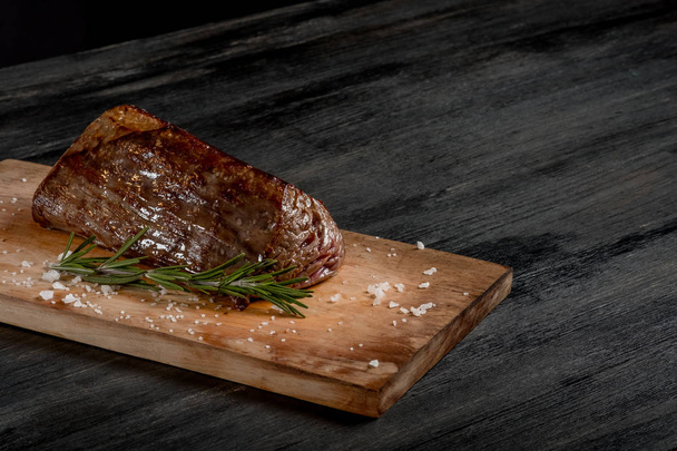 gegrilltes Fleisch mit Gewürzen und Rosmarin auf Holzbrett auf schwarzem Tischhintergrund    - Foto, Bild