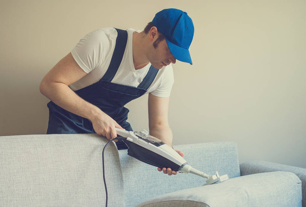 Man in uniform cleaning sofa with dry steam cleaner. Place for text. - Φωτογραφία, εικόνα