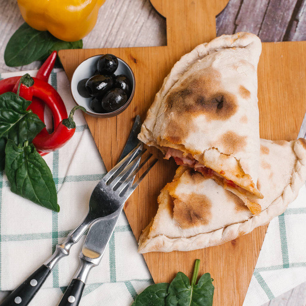 Pizza calzone with decorated edge served on triangular wooden board with knife, fork, basil leaves, red chilli peppers, yellow pepper and olives - Photo, Image