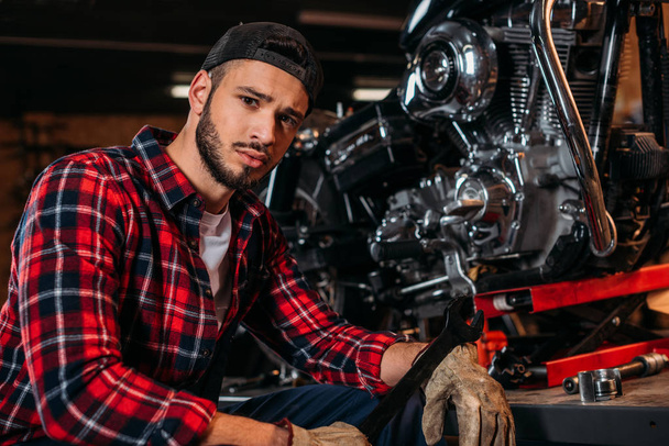 handsome bike repair station worker with wrench sitting in front of motorcycle and looking at camera - Фото, изображение