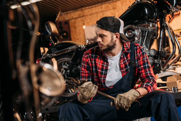 handsome bike repair station worker with wrench sitting in front of motorcycle - Photo, Image