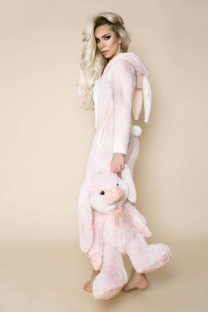 Mooie sexy blonde vrouw die het dragen van een pyjama, een bunny kostuum, lacht gelukkig. Fotomodel op een beige achtergrond in Easter bunny kostuum. - Foto, afbeelding