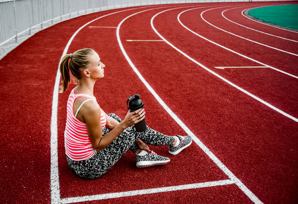 Blonde woman sitting on runnng track and drinking water from  plastic shaker. - Foto, Imagem