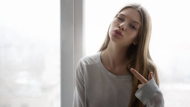 attractive teenager winking and looking at camera while sitting near window  - Фото, зображення