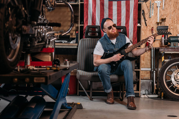 bearded man playing electric guitar at garage with usa flag hanging on wall - Zdjęcie, obraz