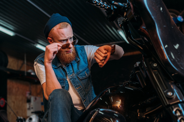 joven guapo en gafas de sol mirando reloj mientras está sentado en la motocicleta en el garaje
 - Foto, Imagen