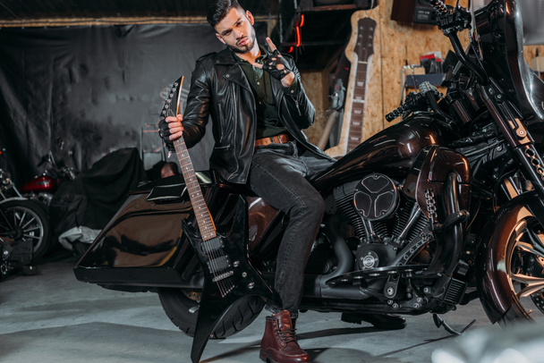 stylish young man in leather jacket sitting on bike with electric guitar at garage - Fotó, kép