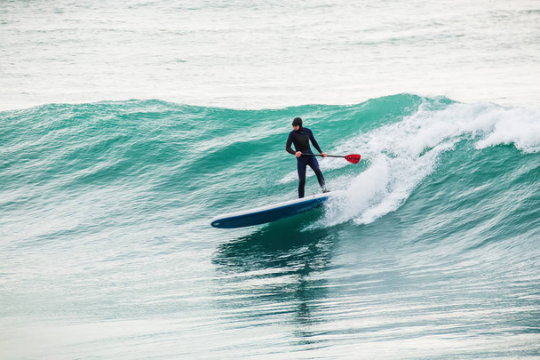 Surfer on stand up paddle board on blue wave. Winter surfing in ocean - Photo, Image